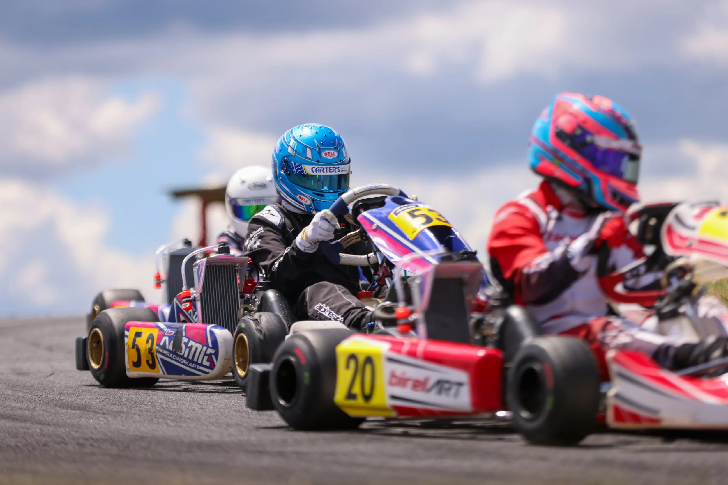 A pack of karts running nose-to-tail through a corner, led by a driver in a blue helmet.